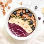 Smoothie Bowl in white bowl topped with granola, banana chips, blueberries and pepitas. Styled with gold spoon and pink linen napkin.
