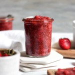 A glass jar of strawberry jam with fresh strawberries.