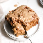 A square slice of apple coffee cake with maple glaze on a small white plate with a fork.