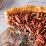 A slice of pecan pie with chocolate chunks in a glass pie dish with metal pie server.