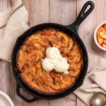 A cast iron skillet of stone fruit cobbler topped with scoops of vanilla ice cream.