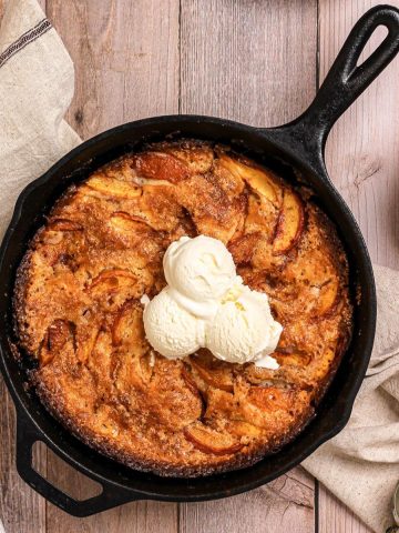 A cast iron skillet of stone fruit cobbler topped with scoops of vanilla ice cream.