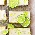 Key lime pie bars with fresh lime slices and lime zest on brown parchment paper.