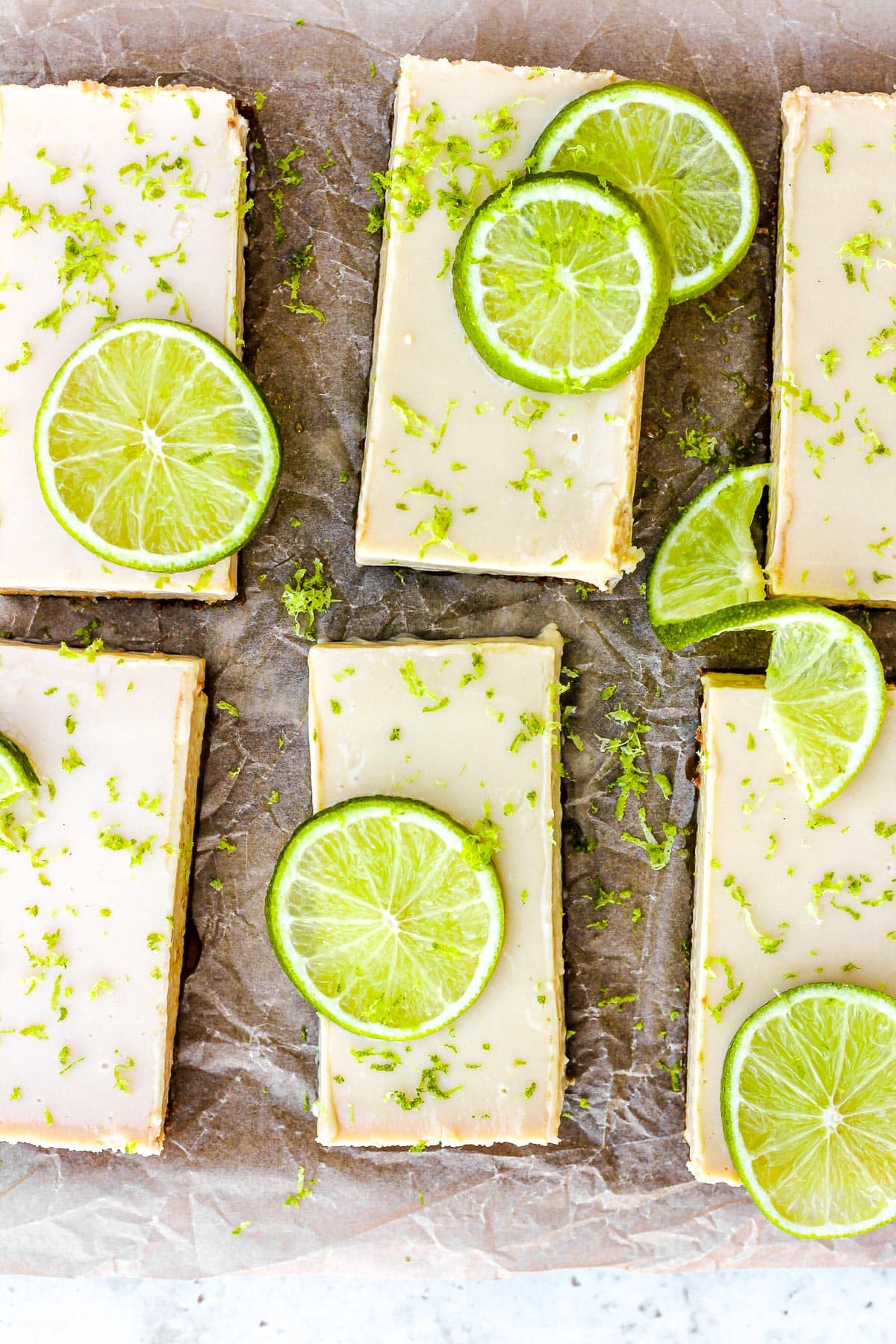 Key lime pie bars with fresh lime slices and lime zest on brown parchment paper.